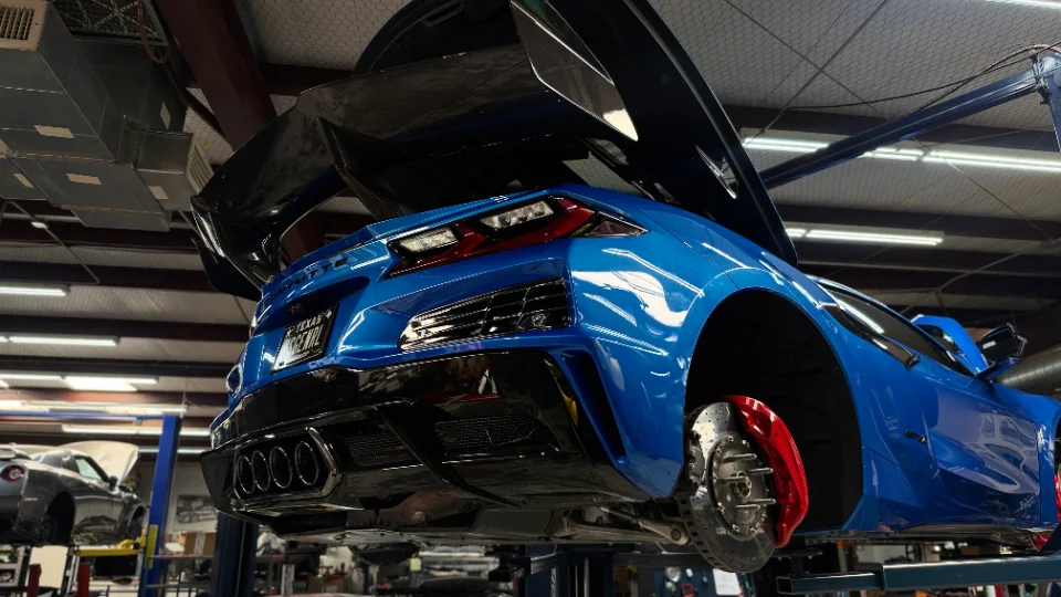 A blue C8 Corvette on a lift in a performance shop, with its wheels removed, showcasing red brake calipers and a large rear wing.