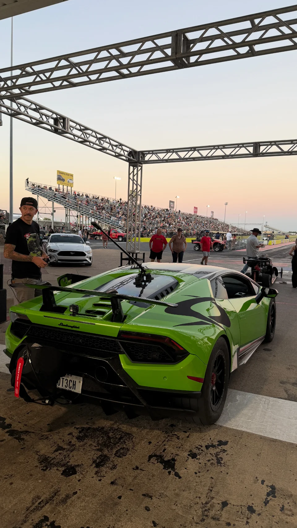 A bright green Lamborghini Huracan with black accents parked at a drag strip, with a crowd of spectators in the background at dusk.