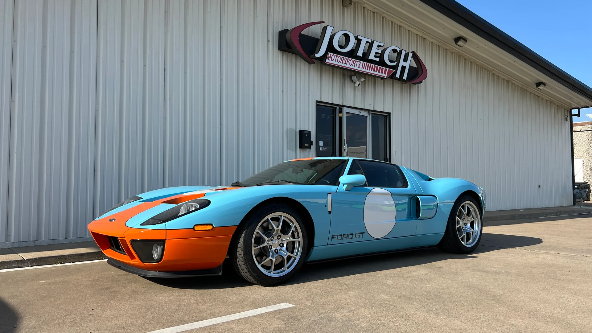 A Ford GT in iconic Gulf livery parked outside the Jotech Motorsports facility on a sunny day.