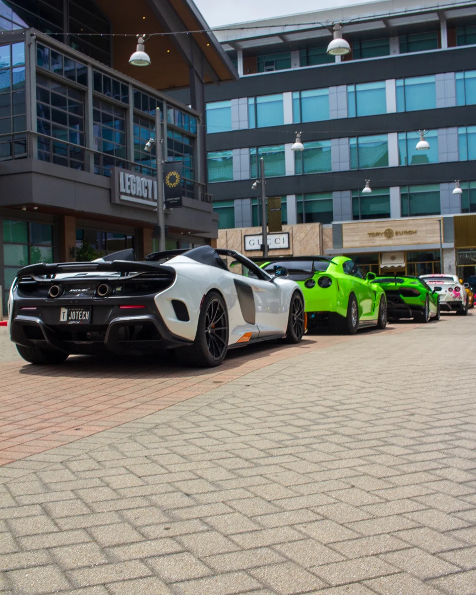 A white McLaren, green Nissan GT-R, and green Lamborghini parked in a row on a brick street in front of shops.