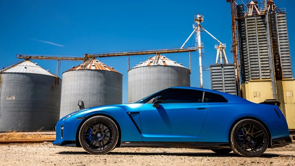 A blue Nissan GT-R R35 with aftermarket wheels parked in front of industrial silos under a clear blue sky.