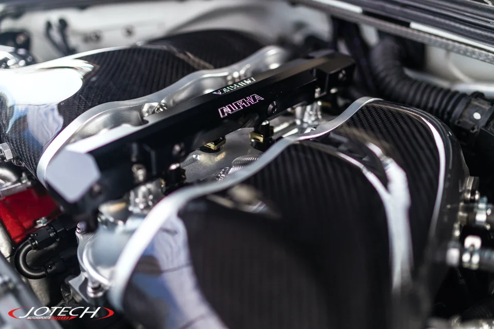 A close-up of a high-performance engine bay featuring carbon fiber intake manifolds and an Alpha Performance fuel rail.