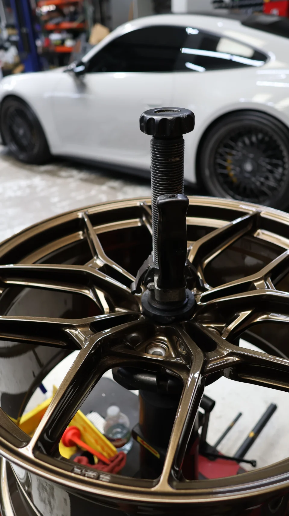 A bronze multi-spoke wheel being mounted on a tire machine in a performance shop, with a white Porsche in the background.