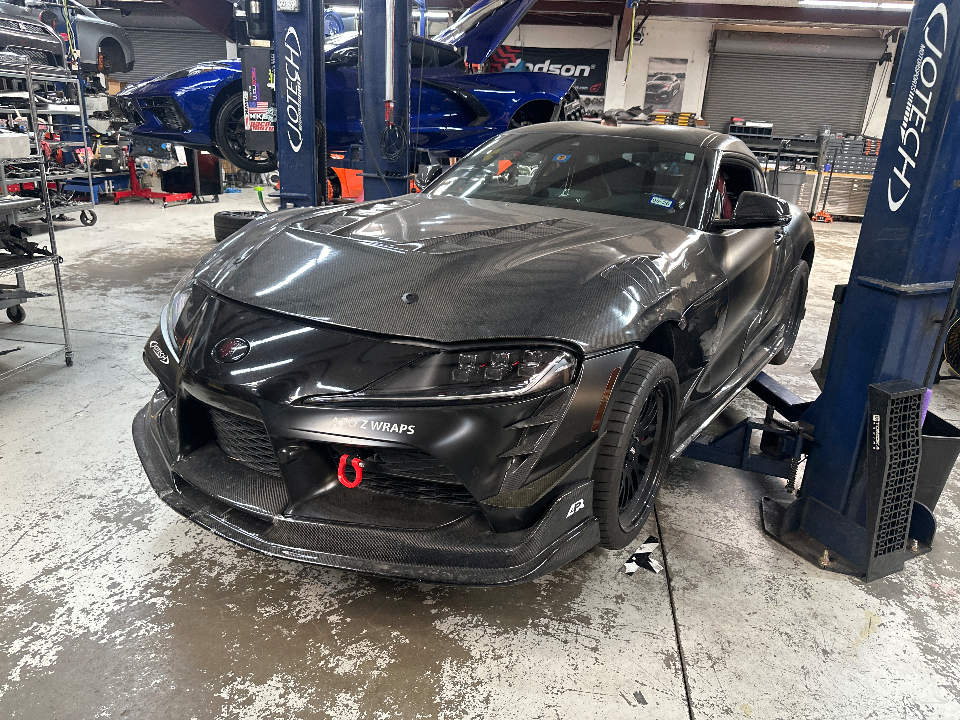 A black carbon fiber Toyota Supra on a lift in a Jotech Motorsports shop, with other cars in the background.