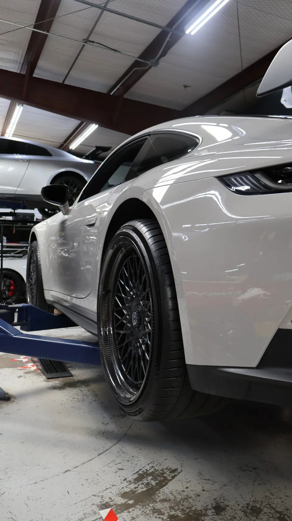 A white Porsche 911 on a lift in a shop, showcasing its black HRE carbon fiber wheels.