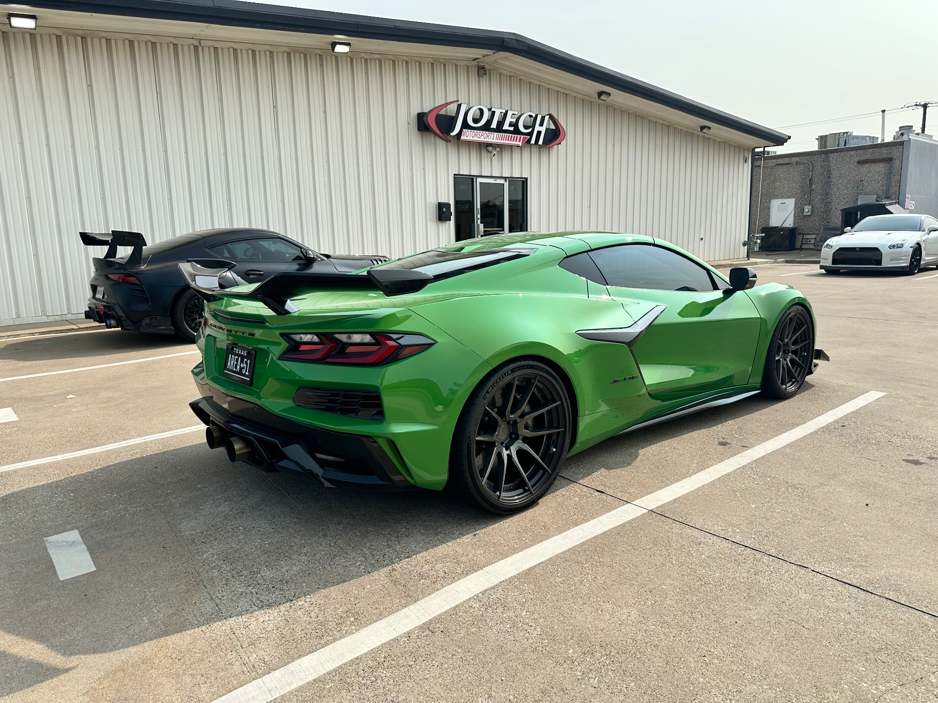 A green C8 Corvette, black Supra, and white GT-R parked outside the Jotech Motorsports facility.
