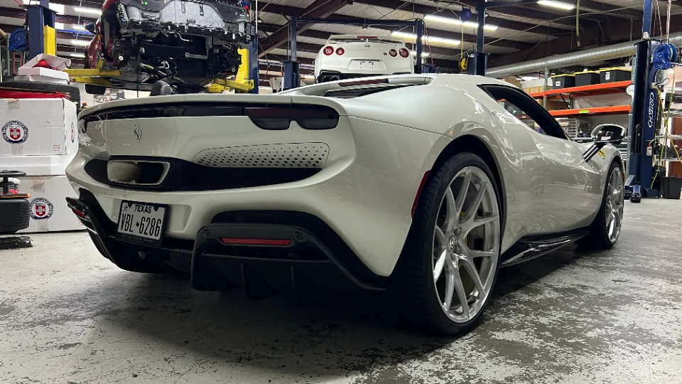 A white Ferrari 296 GTB with aftermarket HRE wheels in a performance tuning shop, with other cars on lifts.