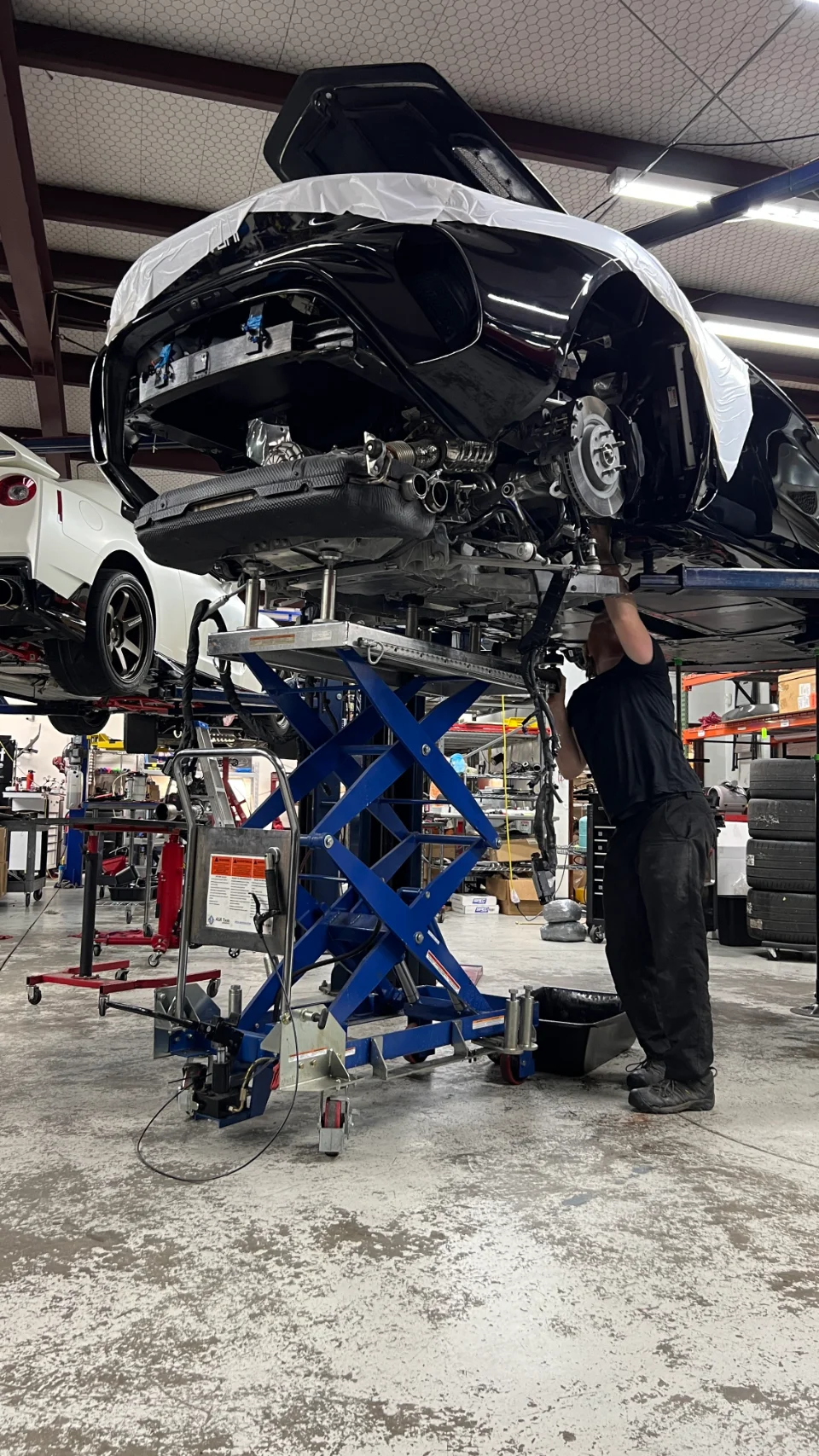 A mechanic works on the exposed rear exhaust system of a black McLaren supercar on a scissor lift in a Jotech Motorsports shop.