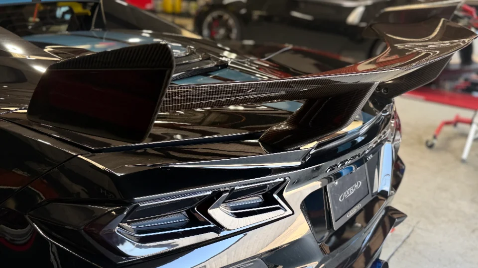 A close-up of the rear of a black Chevrolet Corvette C8, showcasing a large carbon fiber rear wing and LED taillights.