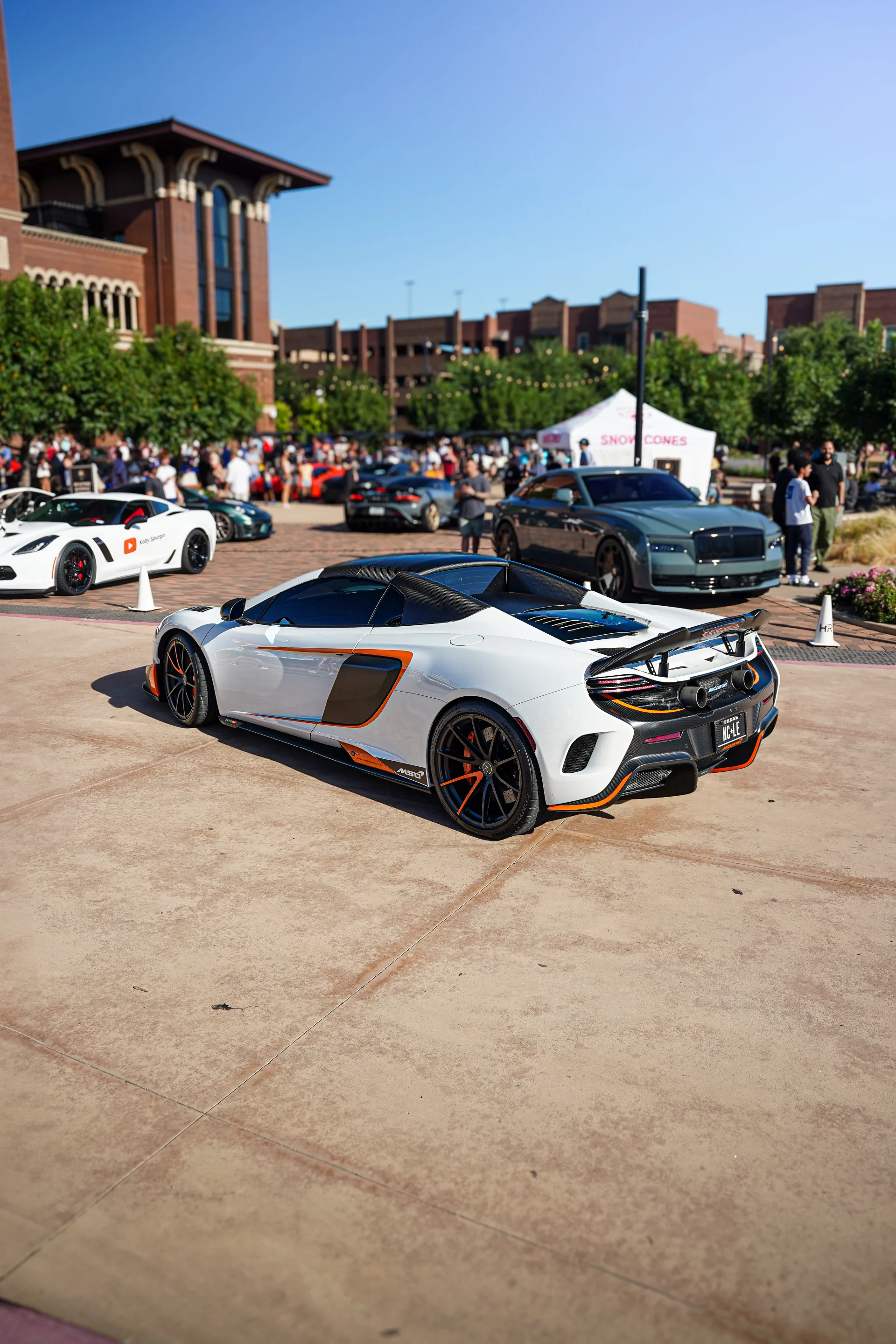 A white McLaren supercar with orange accents parked at an outdoor car show with a crowd and other luxury cars in the background.