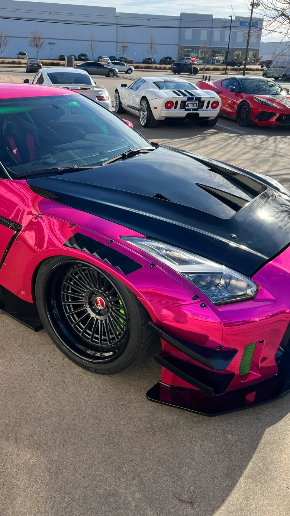 A vibrant pink widebody Nissan GT-R with custom black wheels and a carbon hood, parked outdoors with a white Ford GT and red Corvette C8 in the background.