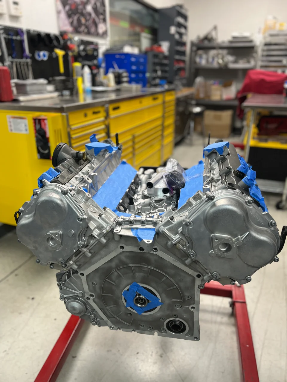 A V8 engine block on an engine stand in a performance tuning shop, with blue painter's tape protecting components during assembly.