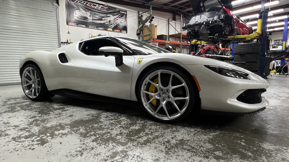 A white Ferrari 296 GTB with aftermarket wheels and yellow calipers parked inside a performance tuning shop.