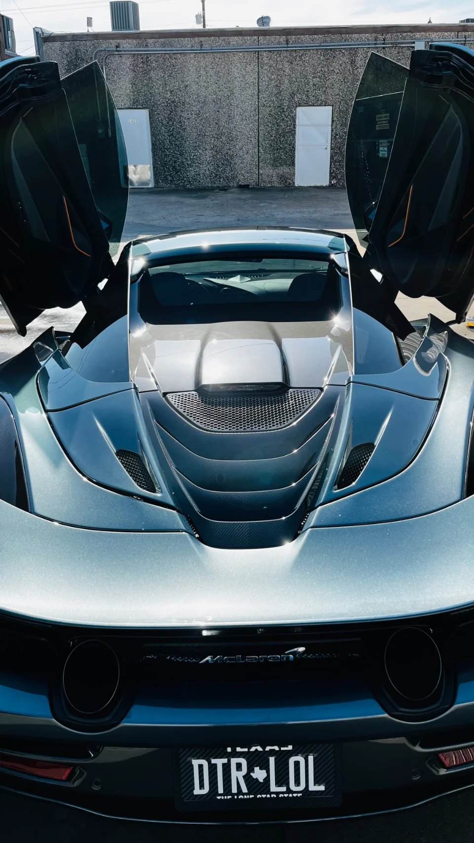 Rear view of a grey McLaren supercar with butterfly doors open, showing exhaust tips and a custom Texas license plate.