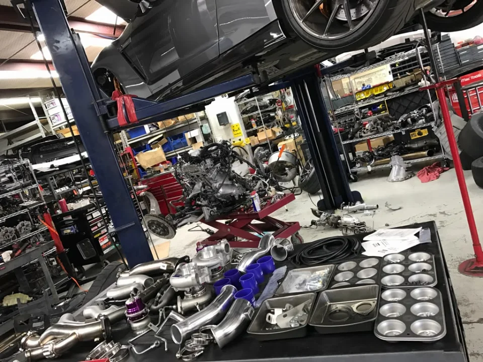 A grey performance car on a lift in a Jotech Motorsports shop, with turbochargers and other performance parts laid out on a table.