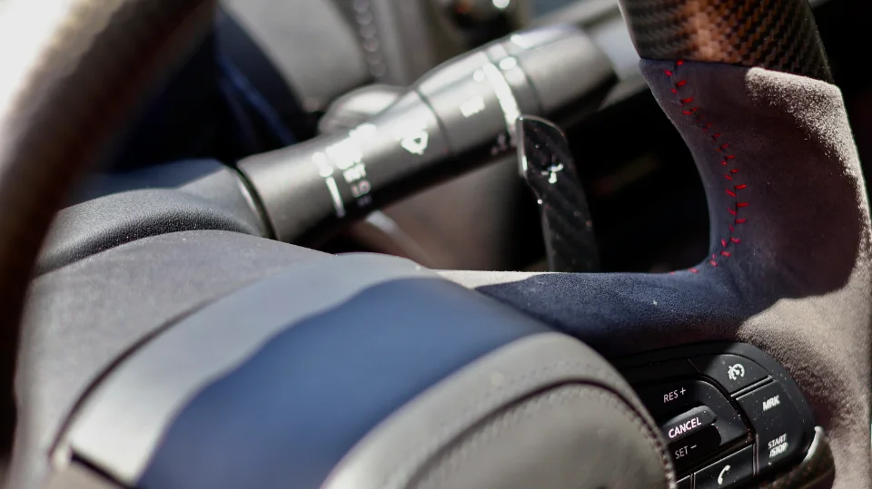 Close-up of a car's interior showing a carbon fiber and Alcantara steering wheel with red stitching and a paddle shifter.