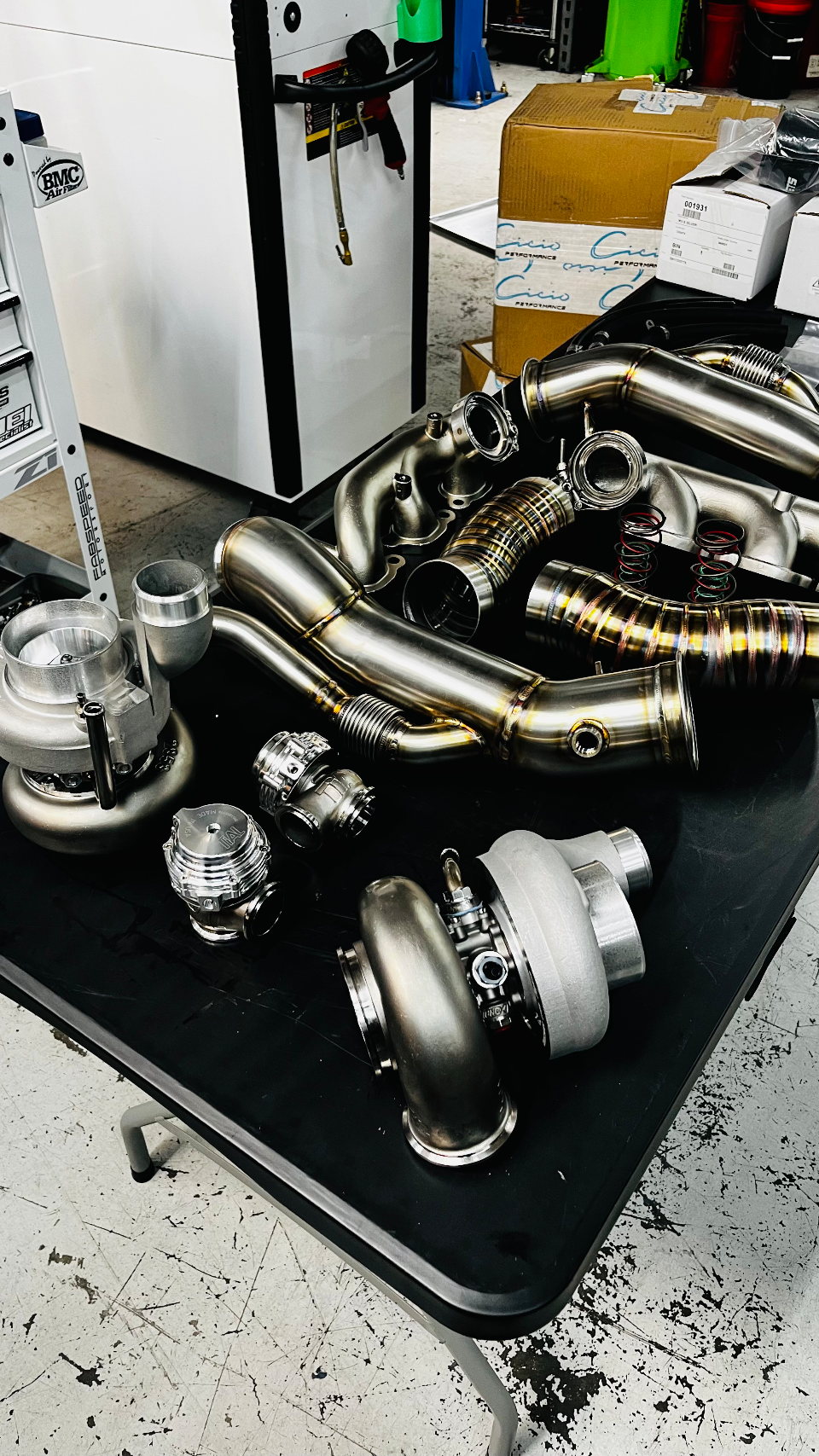 A collection of new stainless steel turbochargers, wastegates, and exhaust piping laid out on a table in a performance shop.