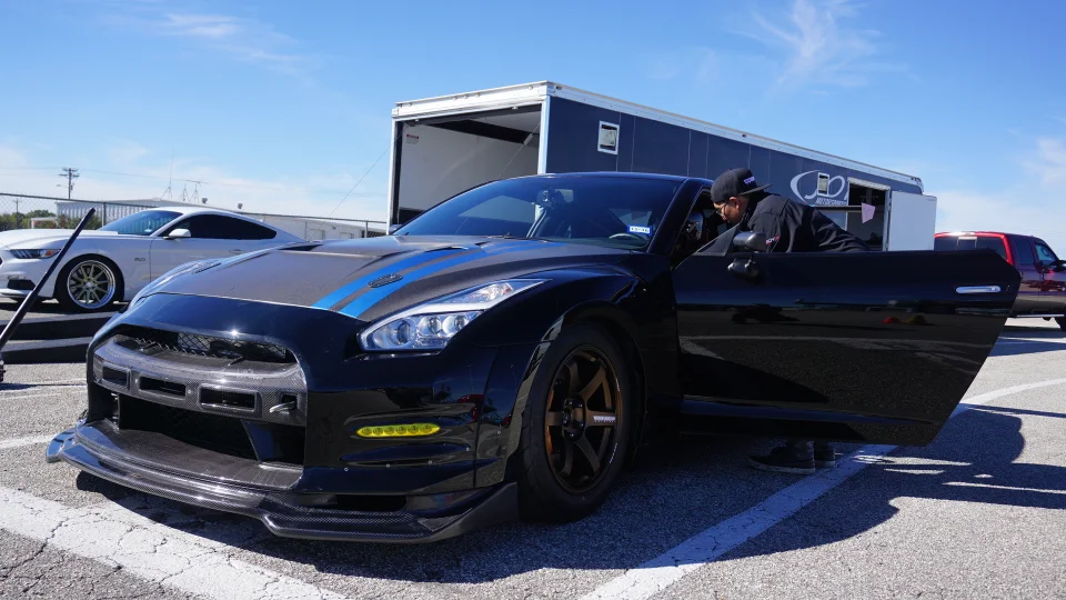 A black Nissan GT-R with carbon fiber aero parts parked at a track event with a white Mustang and trailer in the background.
