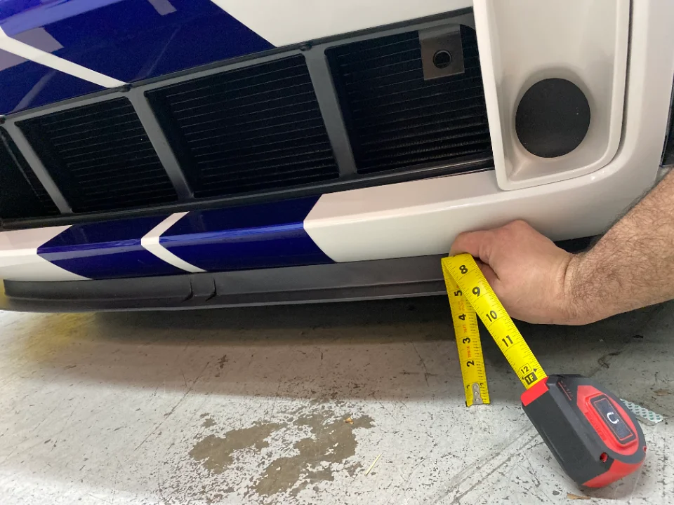 A person measures the ground clearance of a white car with blue racing stripes, showing the front bumper and splitter.