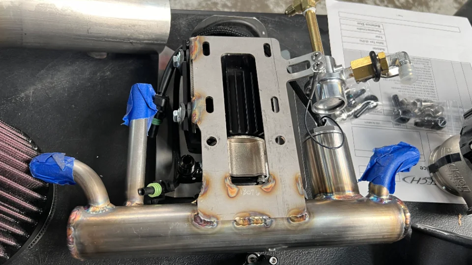 Close-up of a custom stainless steel intake manifold with fuel lines and an air filter, likely for a high-performance build.
