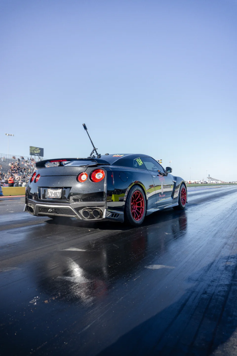 A black Nissan GT-R with red wheels on a drag strip, viewed from the rear, with spectators in the background.