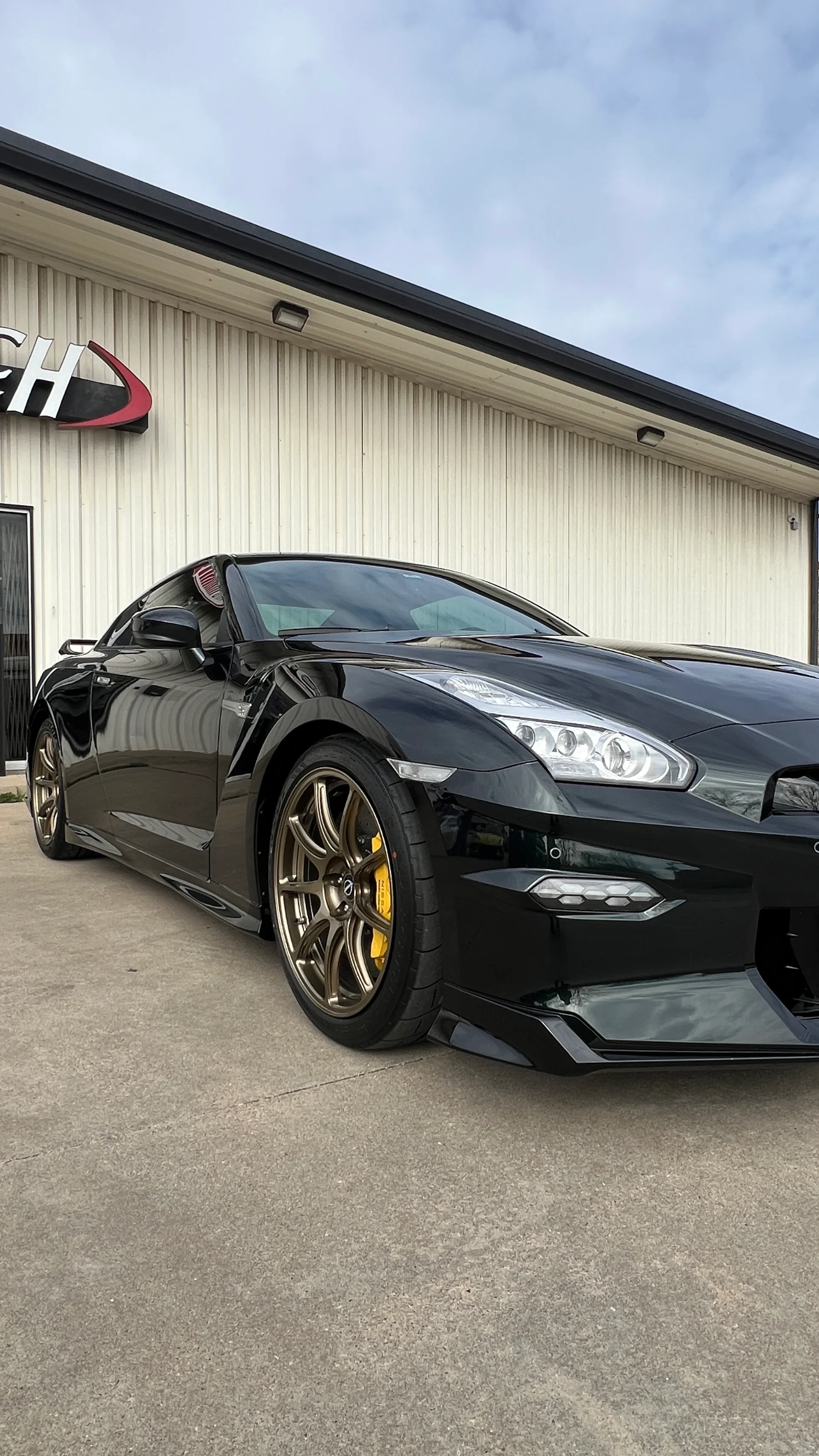 A black Nissan GT-R with bronze aftermarket wheels and yellow brake calipers parked outside the Jotech Motorsports facility.
