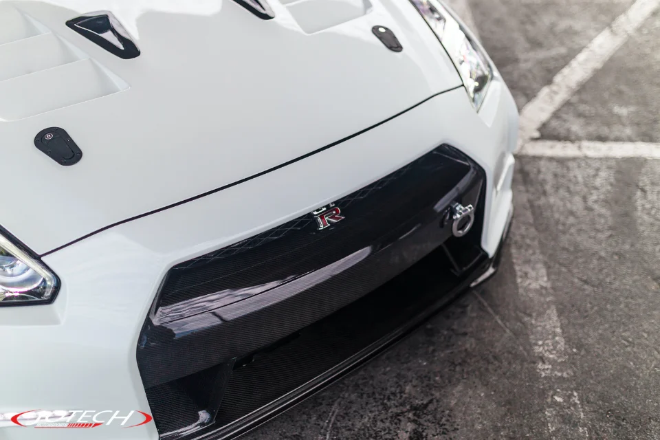 Close-up of a white Nissan GT-R's front end, featuring a carbon fiber grille, hood vents, and hood pins.