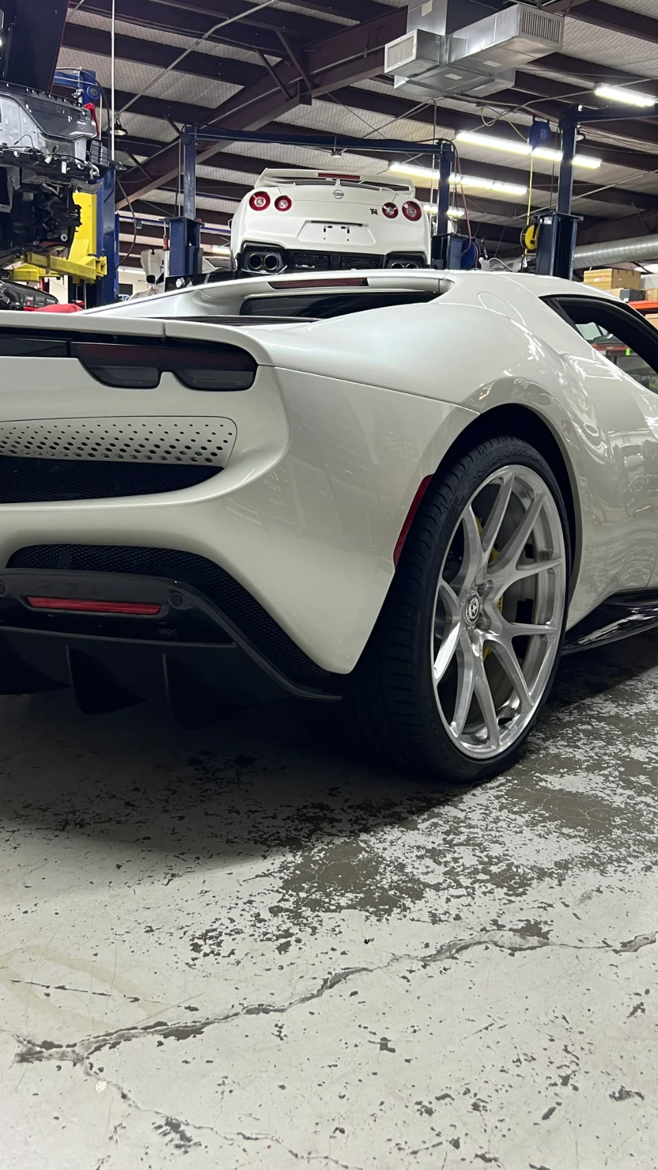 A white Ferrari SF90 with aftermarket wheels in a performance shop, with a white Nissan GT-R on a lift in the background.
