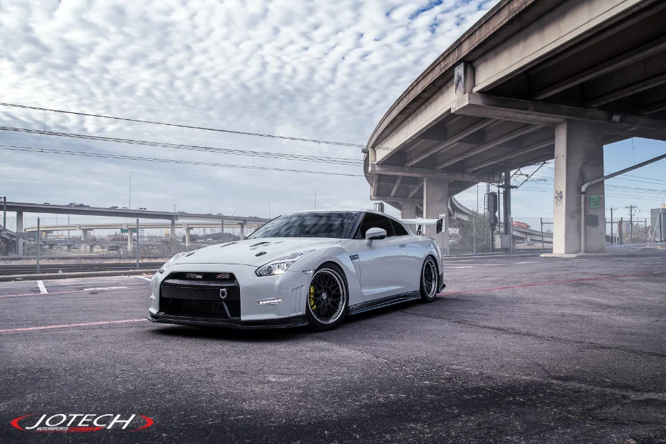 A white Nissan GT-R with aftermarket wheels and a large rear wing is parked under a highway overpass in an urban setting.