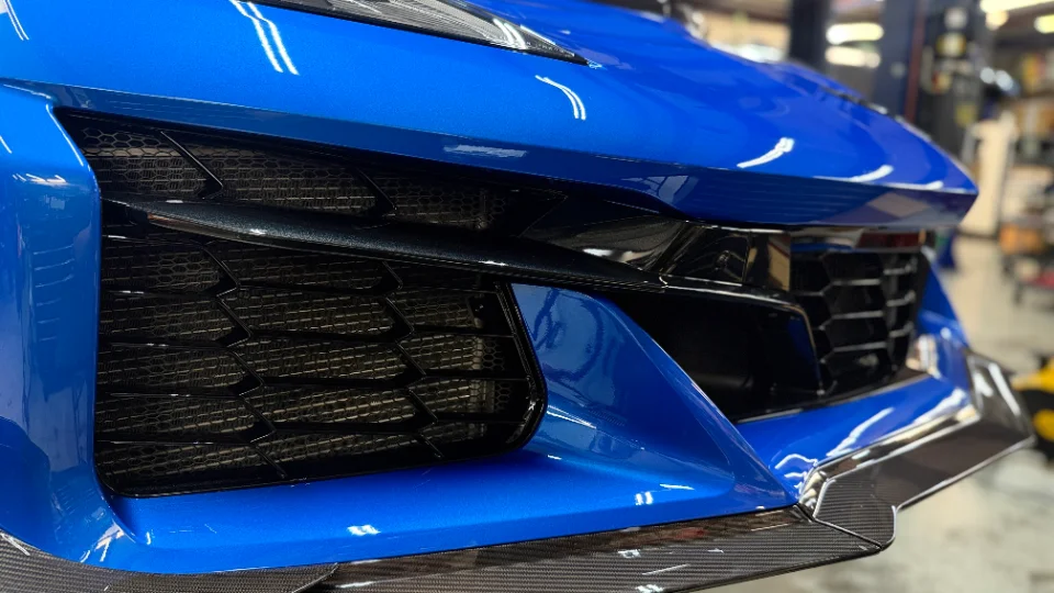 A close-up of the front bumper and grille of a blue Corvette, featuring a carbon fiber splitter.