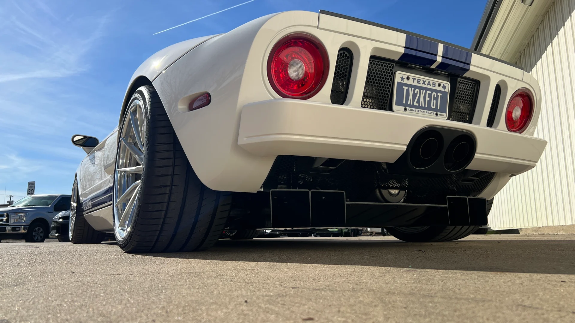 A low-angle rear view of a white Ford GT with blue racing stripes, showcasing its custom exhaust and aftermarket wheels.