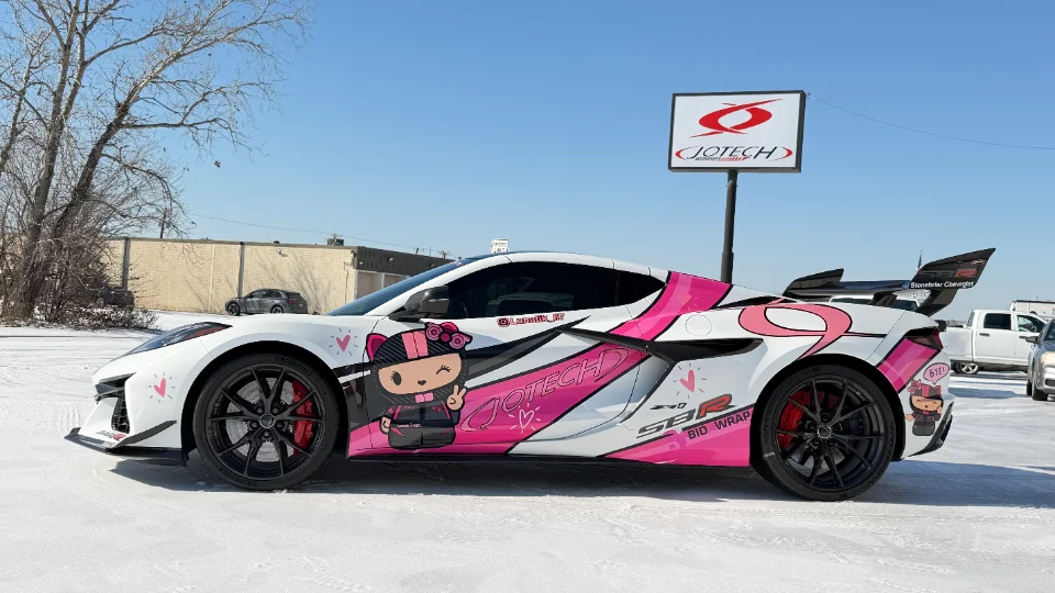 A white Corvette C8 Z06 with a custom pink and black anime-style wrap parked outdoors in front of a Jotech sign.