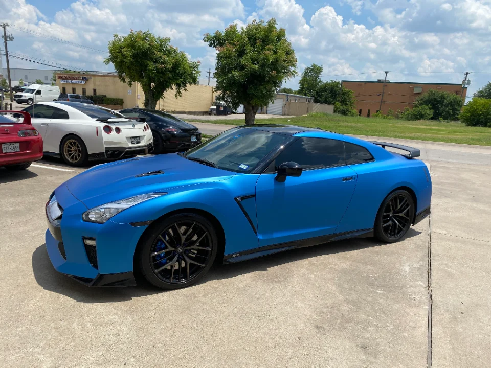 A matte blue Nissan GT-R R35 with black aftermarket wheels parked outdoors, with a white GT-R and red Supra in the background.