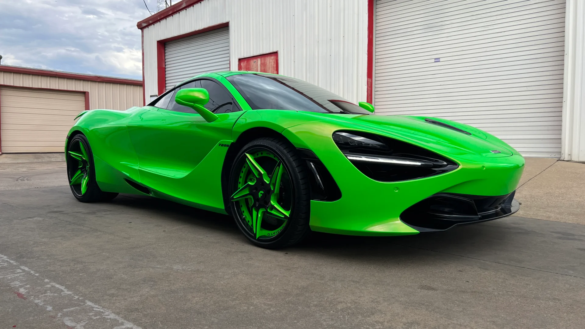 A bright green McLaren 720S with custom green wheels parked in front of a white building with garage doors.