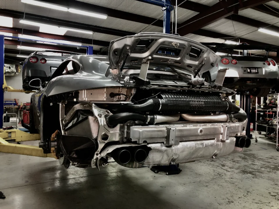 Rear view of a grey Porsche 911 on a lift in a performance shop, with its bumper removed exposing the exhaust system and intercooler.
