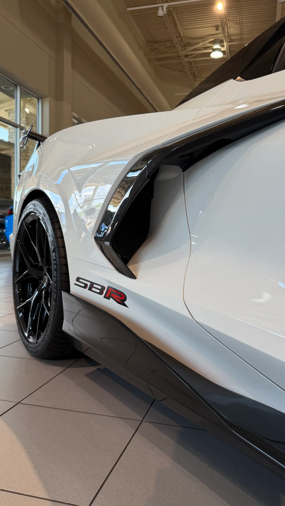 Close-up of a white C8 Corvette's side in a showroom, featuring black wheels, carbon fiber side skirts, and a prominent side air intake.