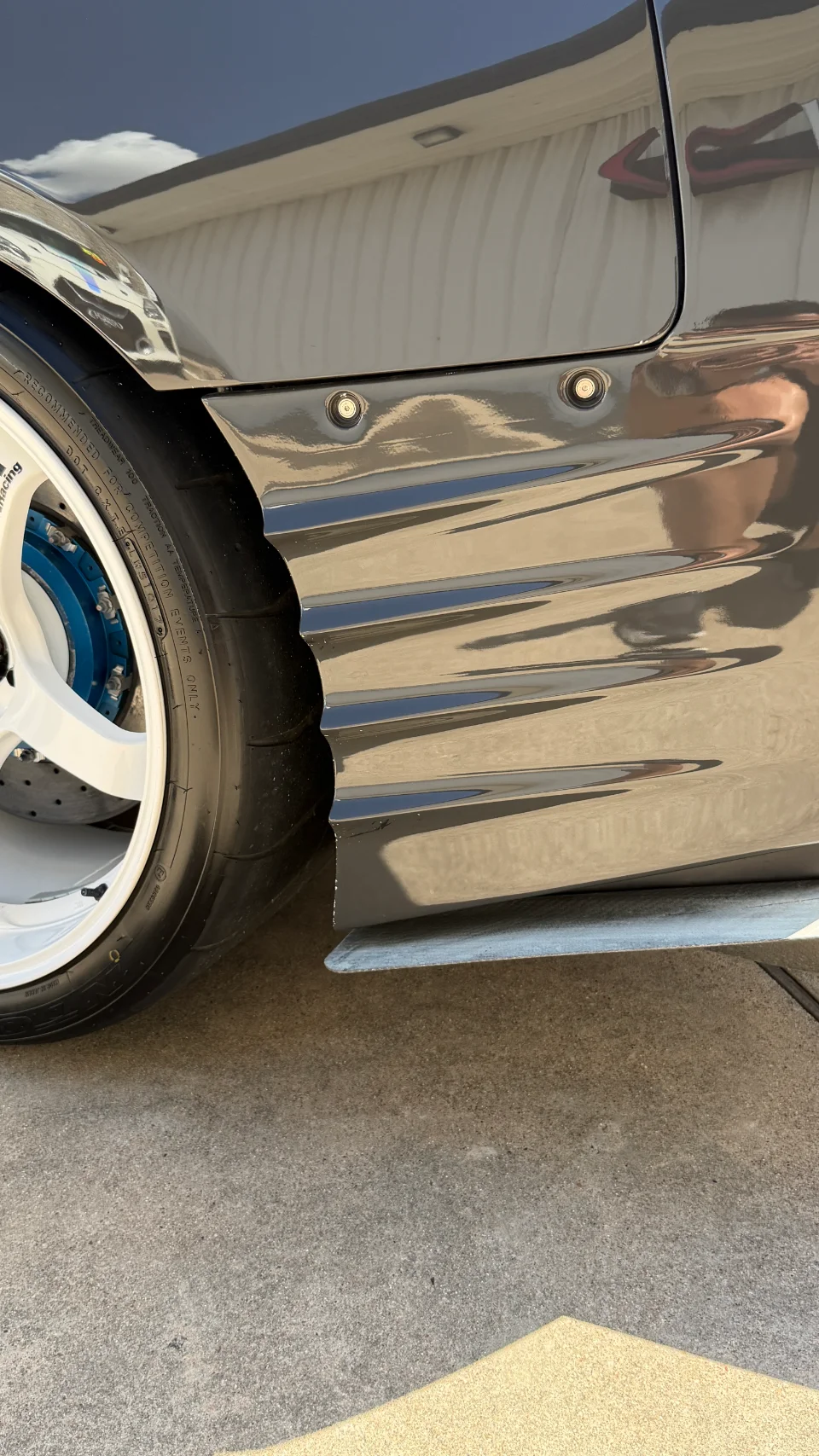 Close-up of a grey car's rear wheel, featuring white multi-spoke wheels, blue brake calipers, and a widebody fender.