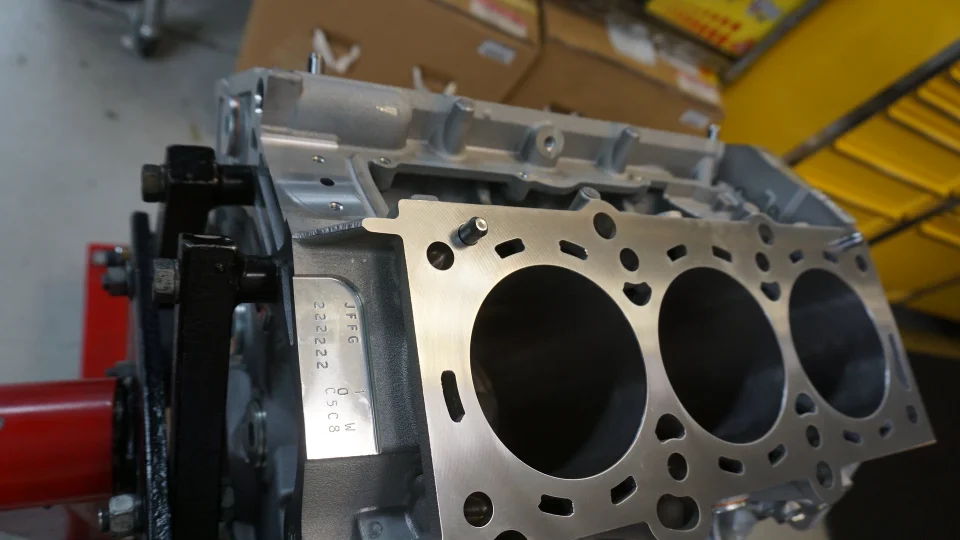 A close-up of an engine block on a stand in a performance shop, showing cylinder bores and the head mating surface.