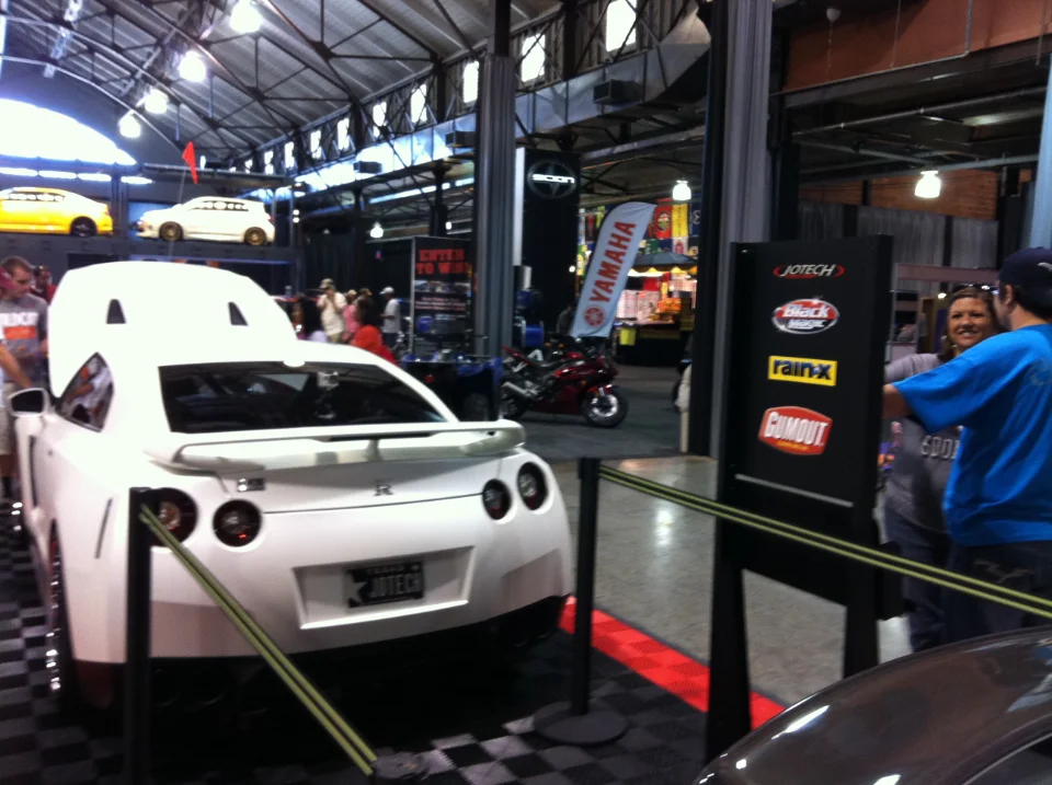 Rear view of a white Nissan GT-R on display at a car show, with vendor booths and people in the background.