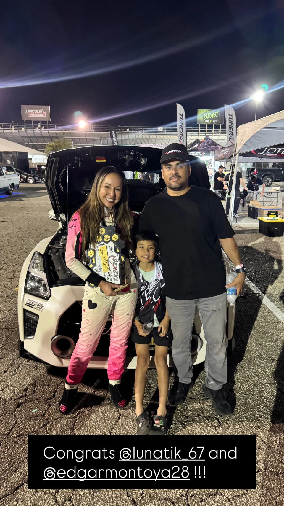 A family celebrates with a trophy next to a white Nissan GT-R at a night drag racing event.