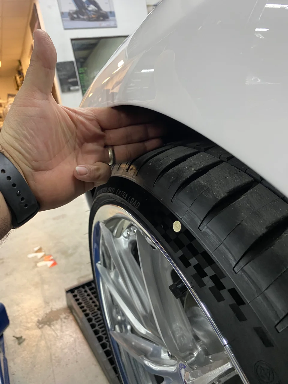 A close-up shot of a hand checking the wheel and tire fitment under the fender of a white car in a shop.