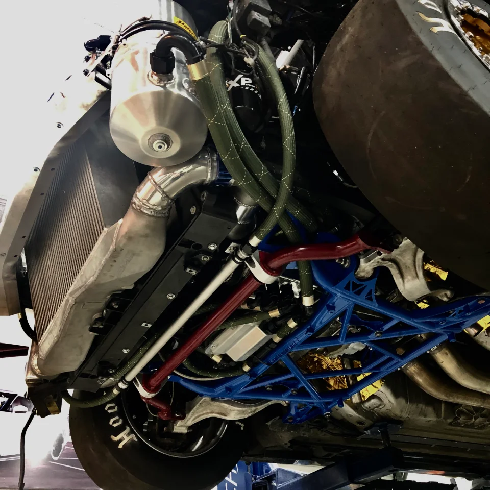 A detailed view of a race car's underbody on a lift, showcasing a large intercooler, custom boost piping, and a blue aftermarket subframe.