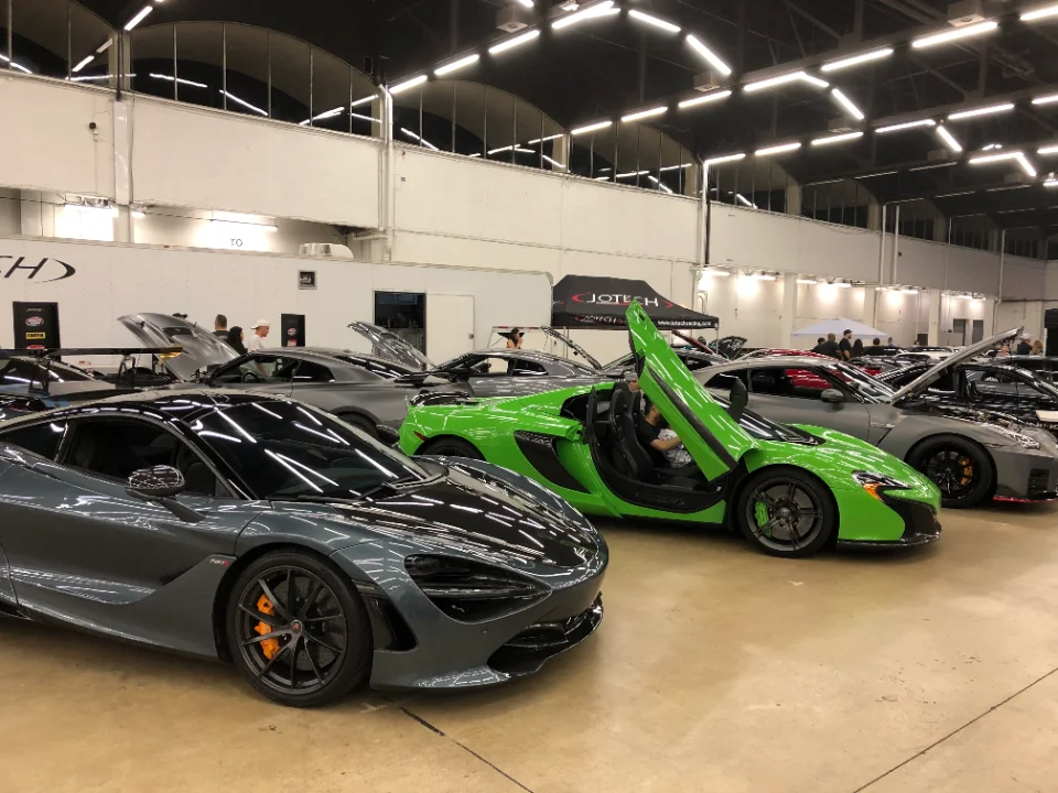 A lineup of high-performance cars including a grey McLaren and a green McLaren with its door open at an indoor car show.