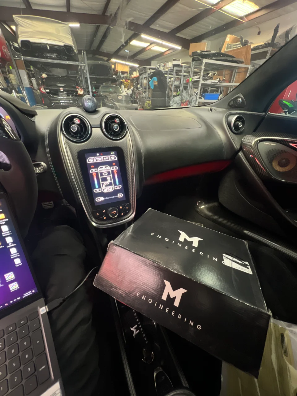 Interior view of a McLaren in a shop with a laptop connected for tuning and an M Engineering product box on the passenger seat.