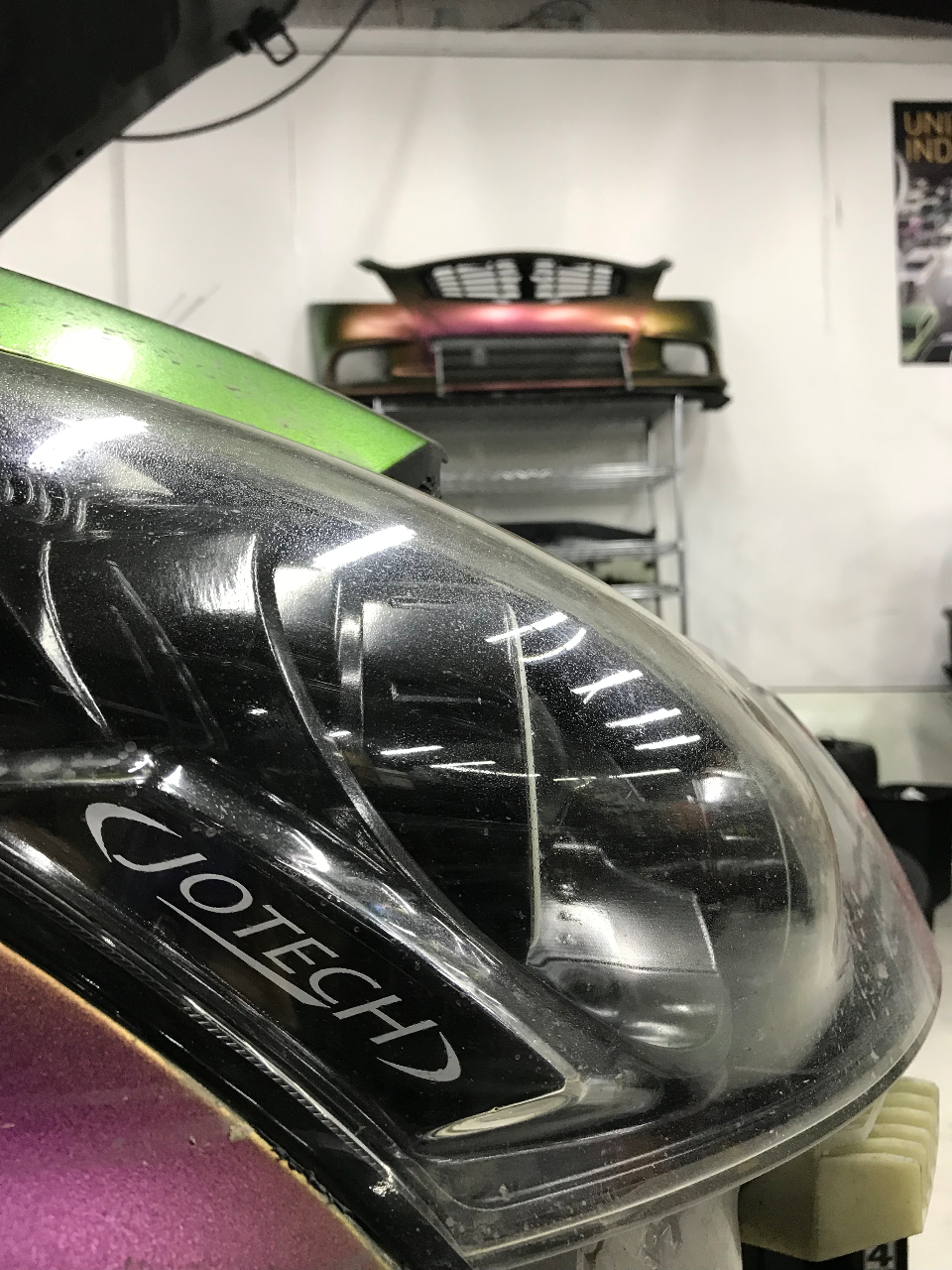 Close-up of a car headlight with Jotech branding, showing iridescent paint on the fender in a workshop setting.