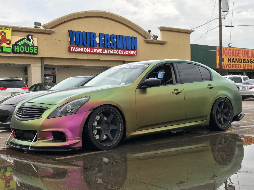 Custom Infiniti G37 sedan with a chameleon wrap, aftermarket wheels, and a front splitter parked on a wet street.