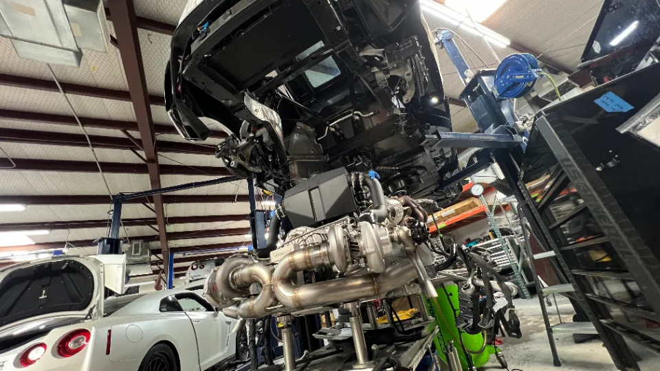 A Nissan GT-R chassis on a lift with its heavily modified twin-turbo engine and custom exhaust system on a stand in a performance shop.