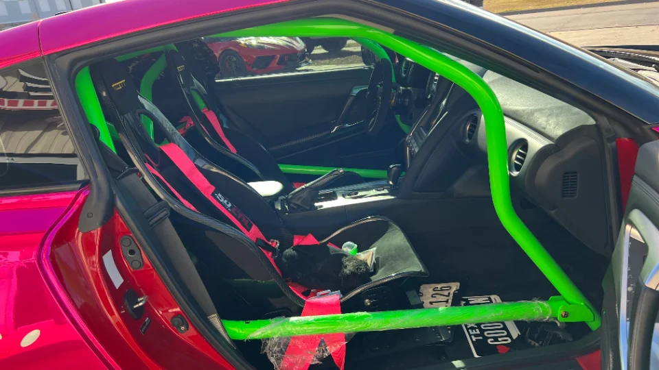 Interior view of a pink Nissan GT-R showing a bright green roll cage, racing seats, and pink harnesses.