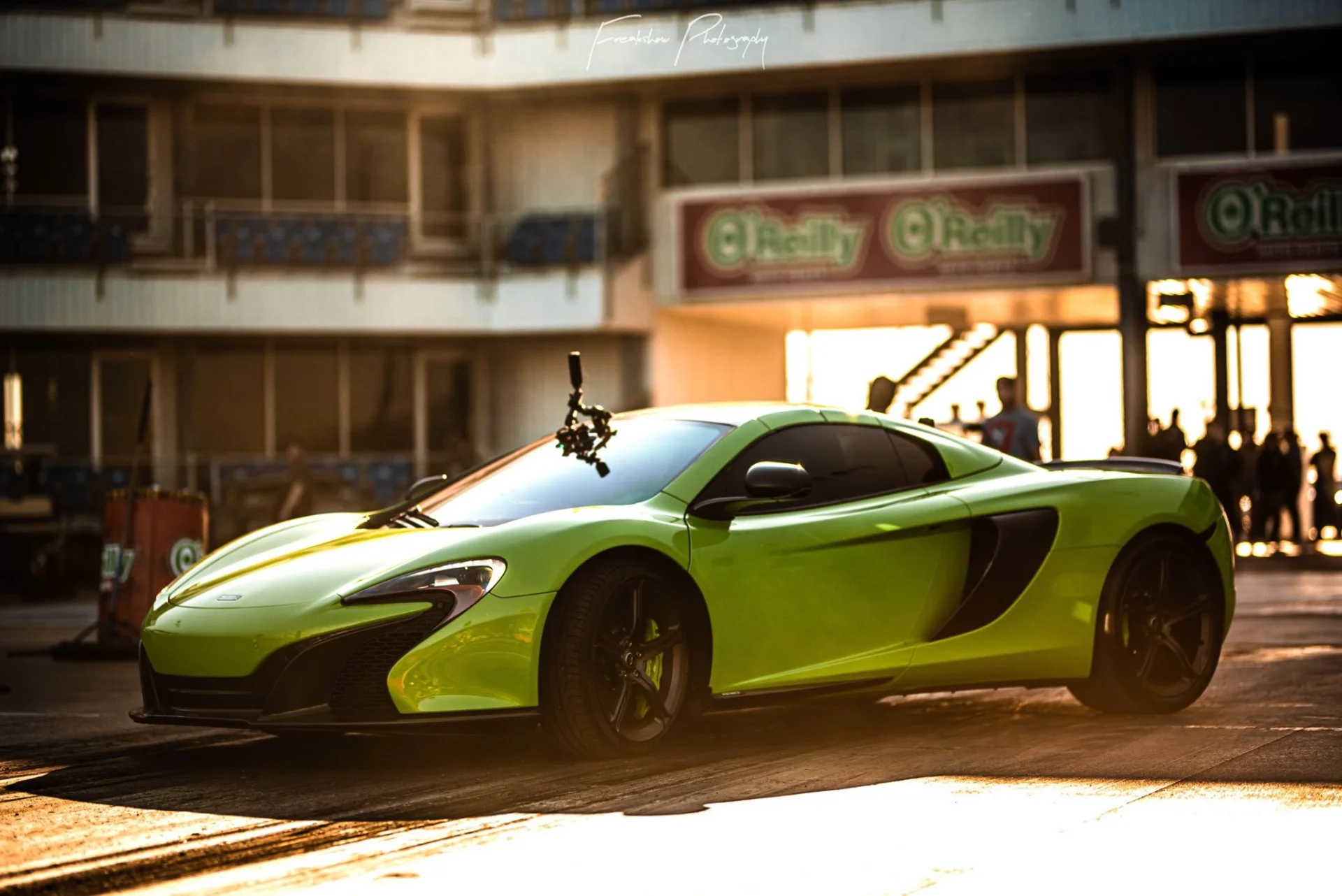 A vibrant green McLaren supercar with a camera rig on its windshield, staged at a drag strip with O'Reilly banners in the background.