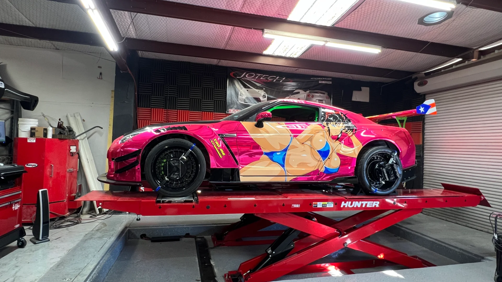 A pink Nissan GT-R with an anime livery on a red shop lift at Jotech Motorsports, ready for service or tuning.